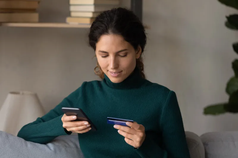 smiling young hispanic latin caucasian woman entering payment information from bank card in mobile shopping app, transferring money, paying for services online, purchasing goods in internet store