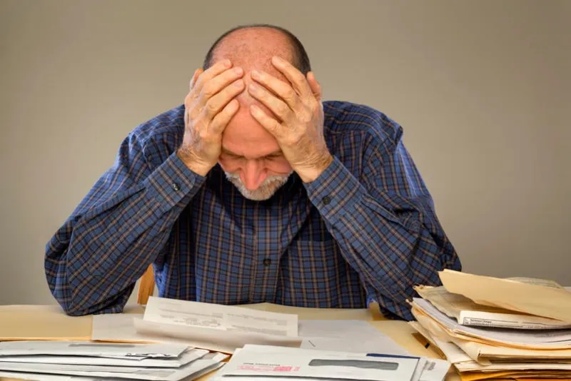 a senior adult man sitting at a table or desk stacked with papers and envelopes looking down with his hands on his head