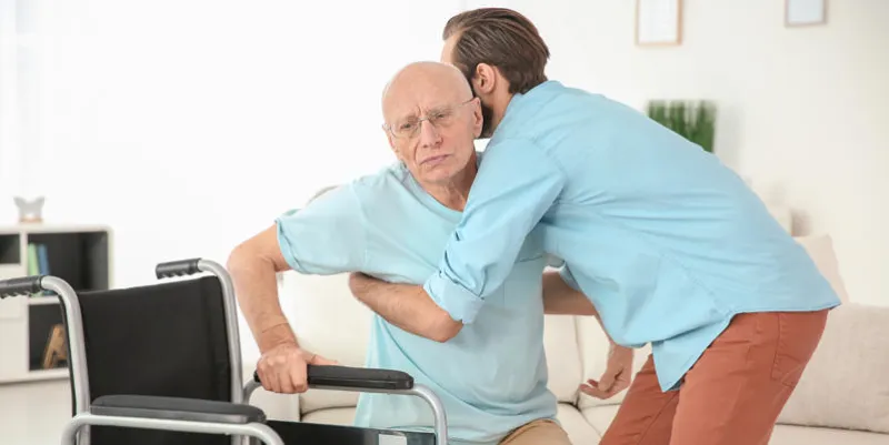 young caregiver helping senior man to sit in wheelchair at home