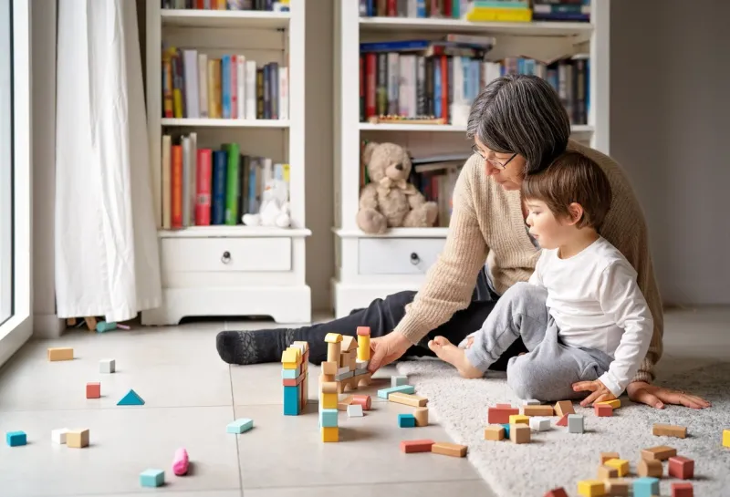 little toddler boy playing wooden building blocks with grandmother at home family spending time together at quarantine period during pandemic bright lifestyle
