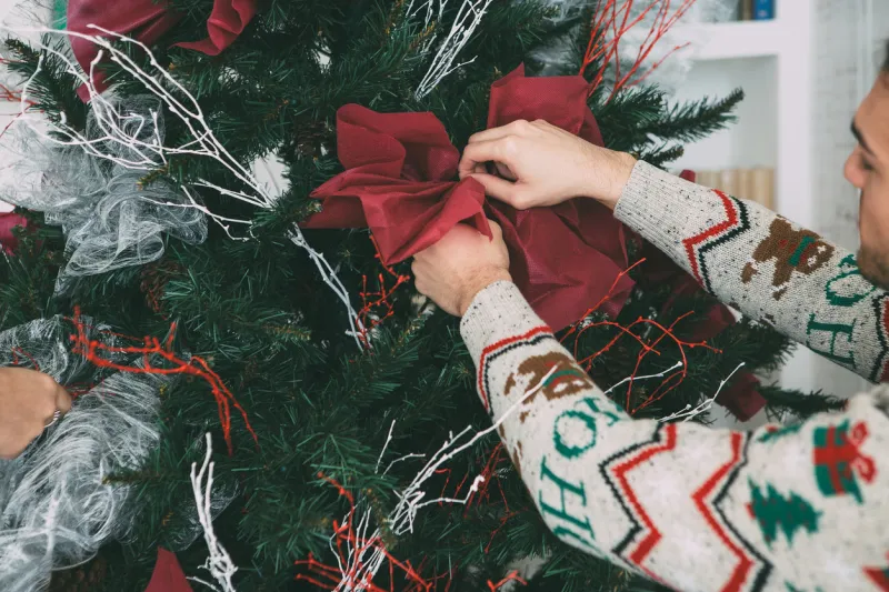 cropped view of a young man and a young woman are decorating a fir tree with christmas ornaments wearing christmas sweaters