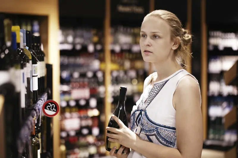 femme achetant du vin ou autre alcool dans un magasin de bouteilles debout devant des étagères pleines de bouteilles avec une expression sérieuse alors qu'elle essaye de se décider