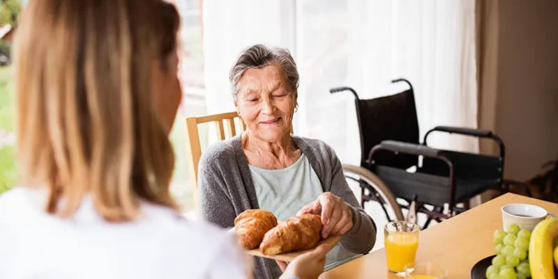health visitor and a senior woman during home visit a nurse giving food to an elderly woman sitting at the table