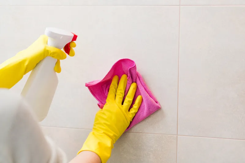 hands with yellow rubber gloves holding detergent spray bottle and cleaning tiles in bathroom with pink cloth spring cleaning