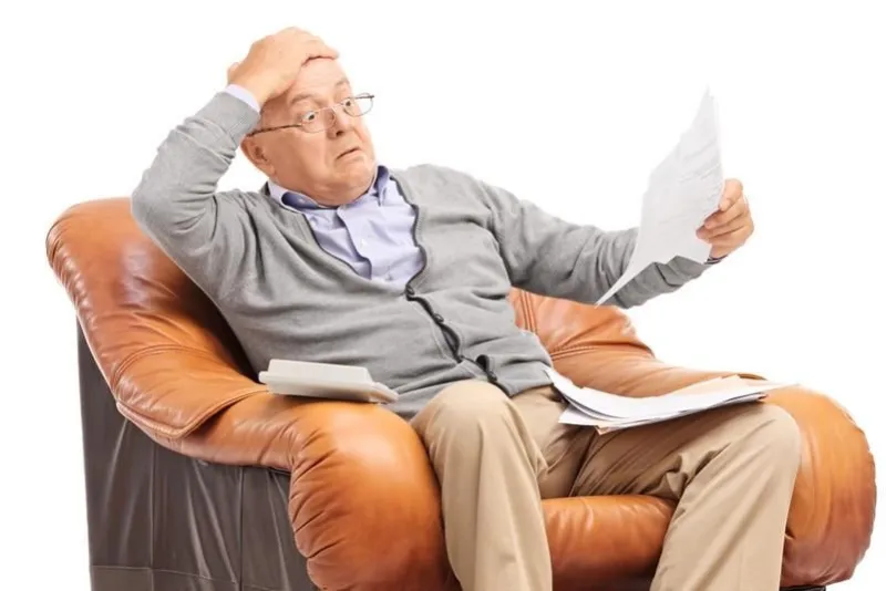 shocked senior gentleman looking at his bills in disbelief seated in an armchair isolated on white background
