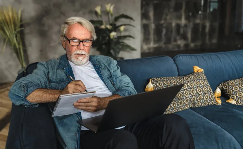 focused elderly man writing notes in notebook watching webinar or online training using laptop computer, modern senior male with gray hair and beard learning online