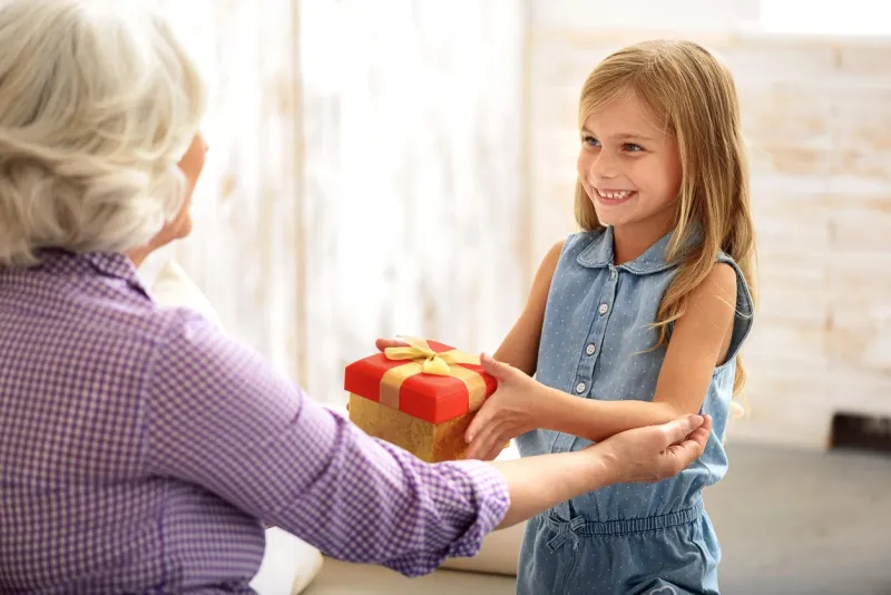 i present is for you joyful little girl is giving box to her granny she is standing and smiling mature woman is embracing her with love