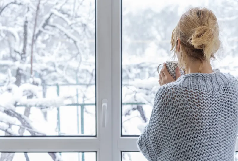 a woman in a cozy sweater stands at the picture window with a cup of drink in her hands