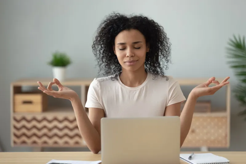 african woman meditating sit at desk in front of pc, serene mixed-race female closed eyes folded fingers mudra symbol do exercise practising yoga reducing anxiety stress positive frame of mind concept