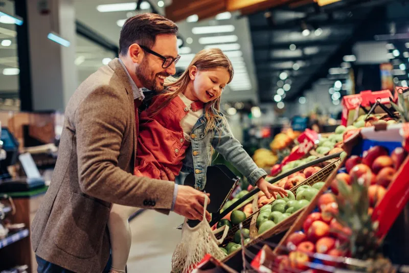 little girl is buying groceries in the supermarket with her father he carrying her and picking together fresh fruits
