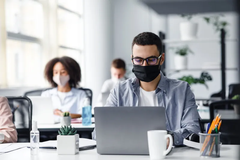 office center workers are protected from virus outbreak during covid-19 epidemic young hipster man in glasses and protective mask works at laptop, with colleagues at workplaces in interior, free space