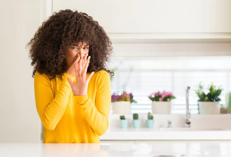 african american woman wearing yellow sweater at kitchen smelling something stinky and disgusting, intolerable smell, holding breath with fingers on nose bad smells concept
