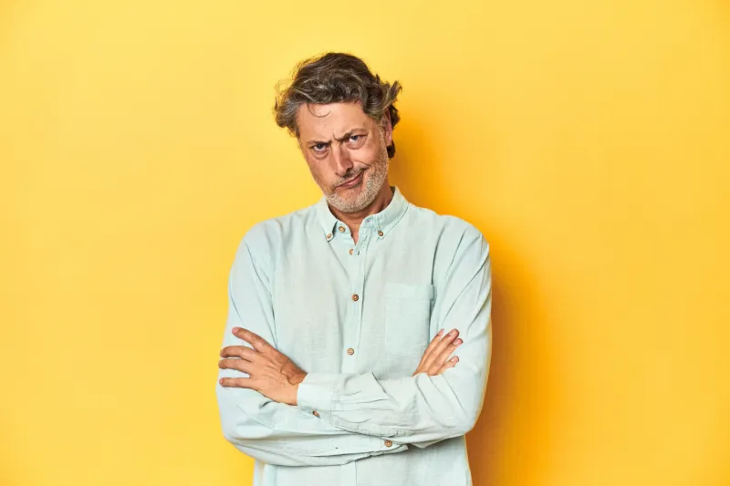 middle-aged man posing on a yellow backdrop frowning face in displeasure, keeps arms folded