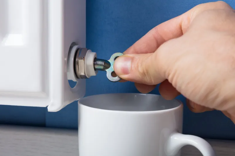 close-up of person's hand turning radiator bleed valve to release air with cup at home