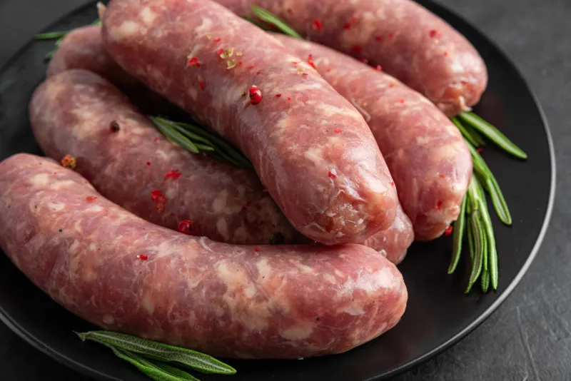raw sausages or bratwurst with spices and rosemary in a plate on black background close up food for grill or barbeque