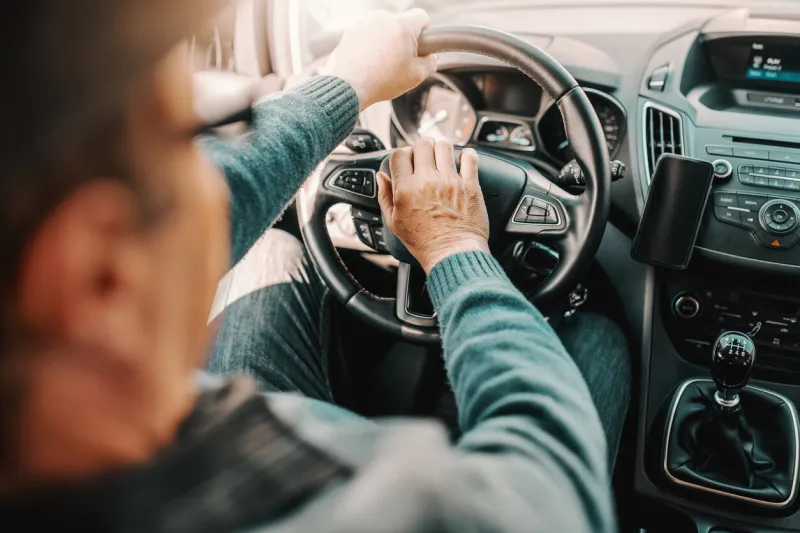 close up of senior man driving his car one hand on steering wheel and other on horn picture taken from back seat