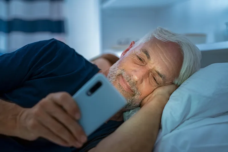 senior man watching video on smart phone while in bed smiling mature man lying on bed using cellphone in the dark at home addict man awake using phone for chatting late in night