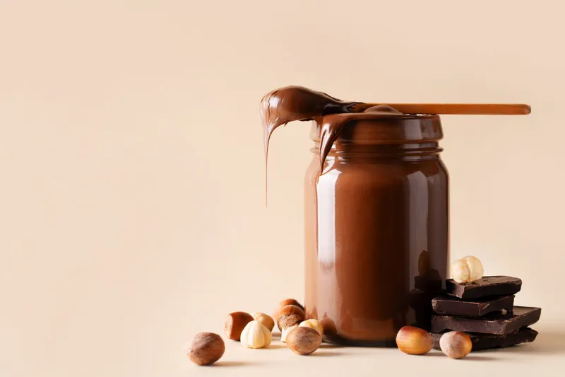 chocolate paste in jar with nuts on cut wooden board on beige background, close up