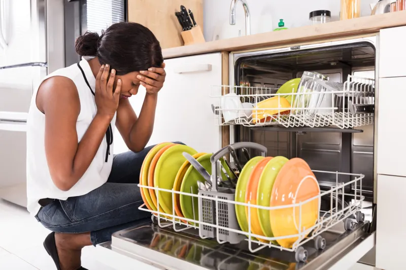sad african woman crouching near dishwasher in kitchen