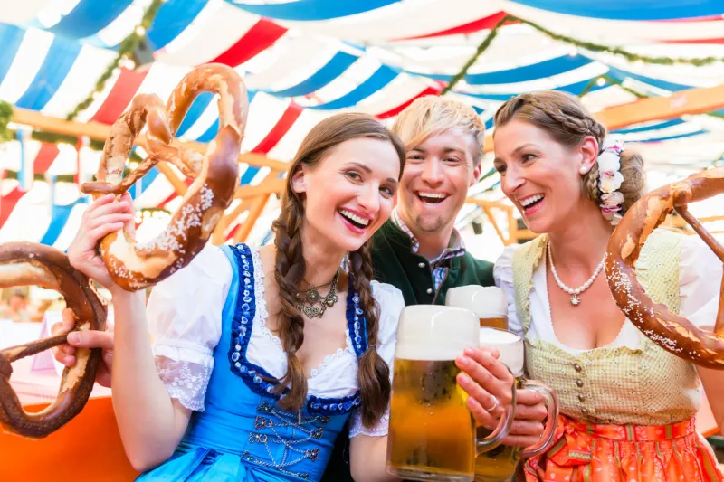 three friends in beer tent at dult or oktoberfest holding giant pretzels up in the air