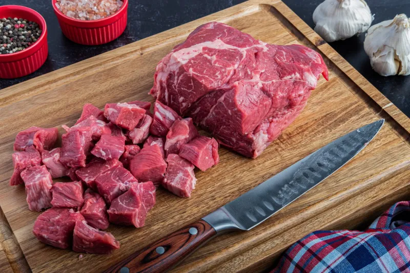 a boneless beef chuck roast being cubed for a stew on a cutting board with sea salt and peppercorns next to a carving knife
