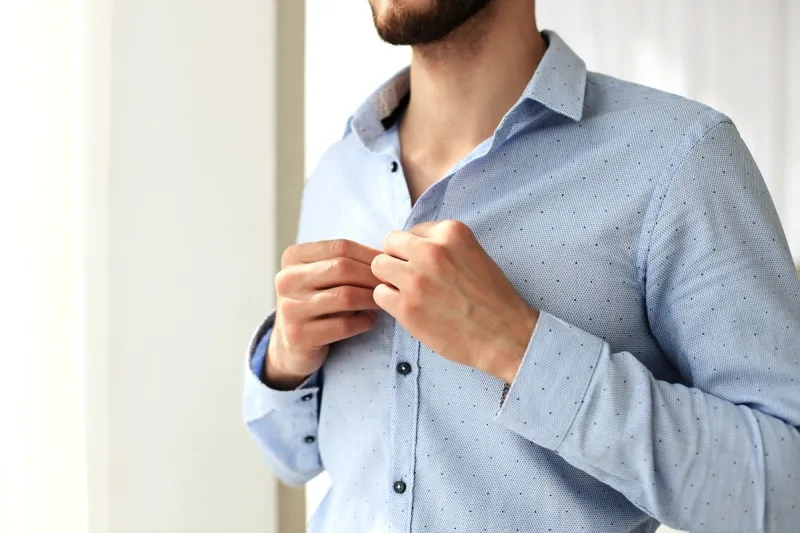 man buttons sleeves on blue linen dress shirt in bedroom at slight angle