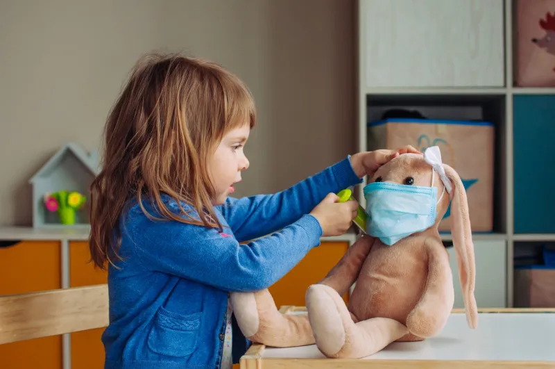 toddler girl playing doctor giving an injection bunny soft toy in the face mask in the kids room pandemic covid-19 concept