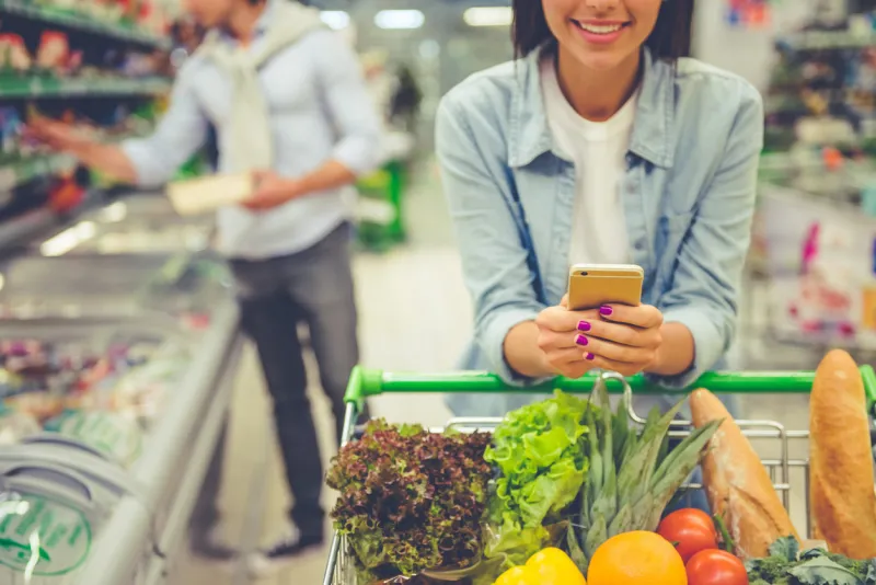 couple in the supermarket cropped image of girl leaning on shopping cart, using a mobile phone and smiling, in the background her boyfriend is choosing food