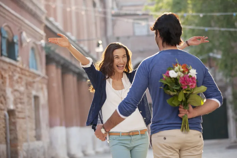 enthusiastic woman approaching man with flowers behind back