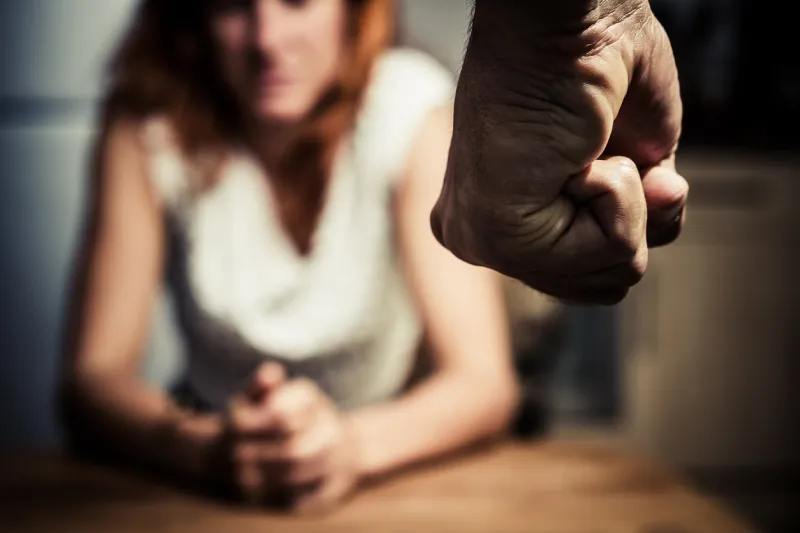 young woman is sitting hunched at a table at home, the focus is on a man's fist in the foregound of the image