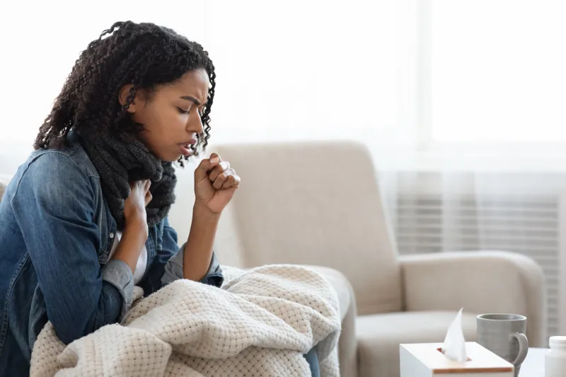 risk of coronavirus sick black woman coughing hard at home, sitting on couch wearing scarf and covered with blanket, side view with copy space