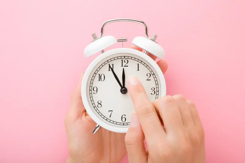 young woman hands holding white alarm clock finger pointing to arrow of twelve o'clock light pink table background pastel color time change concept closeup top down view