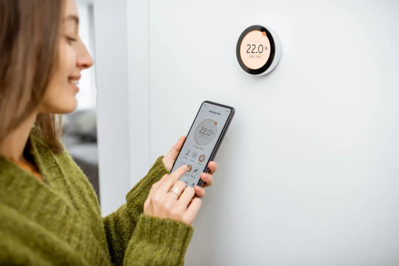 woman dressed in green sweater regulating heating temperature with a modern wireless thermostat and smart phone at home synchronization of thermostat with mobile devices concept