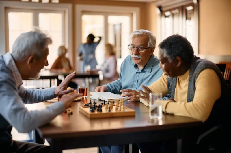 mature man reading book and communicating with male friends while they are playing chess at retirement community