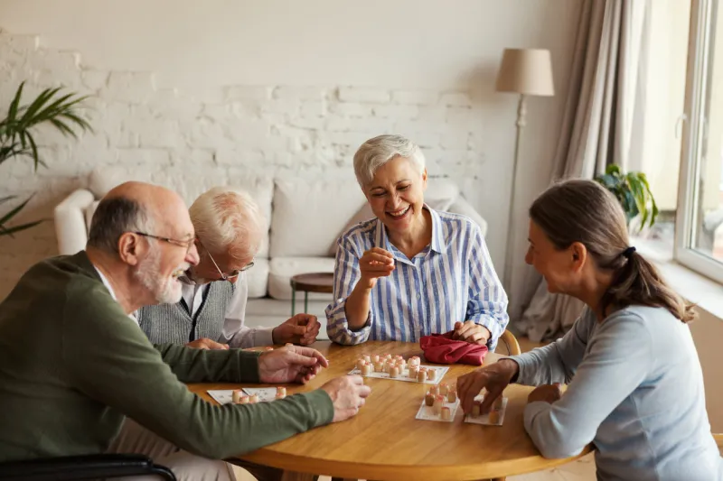 group of four cheerful senior people, two men and two women, having fun sitting at table and playing bingo game in nursing home