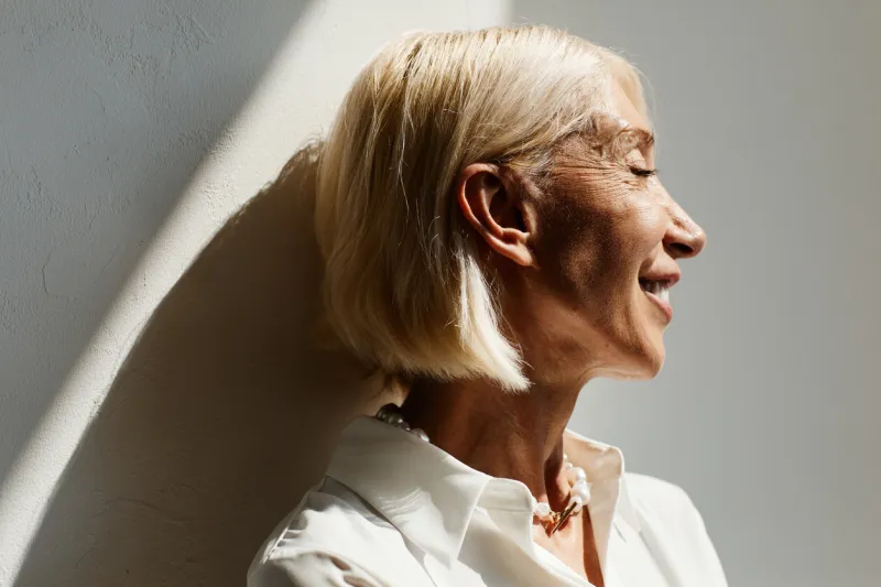 minimal side view portrait of elegant mature woman lit by sunlight against white wall, copy space