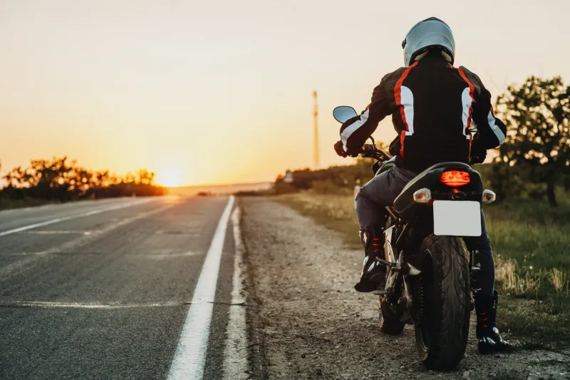 back view of male in protective jacket, boots and helmet riding off roadside at sunset on empty highway backlit background