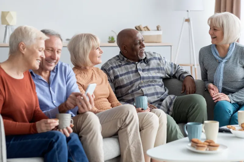 international group of cheerful elderly people men and women in casual outfits chilling together at sanatorium, sitting on sofa in living room, using mobile phone, drinking tea, chatting