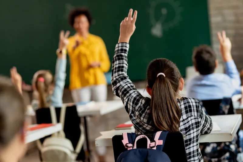 back view of elementary student raising arm in order to answer a questing during a class