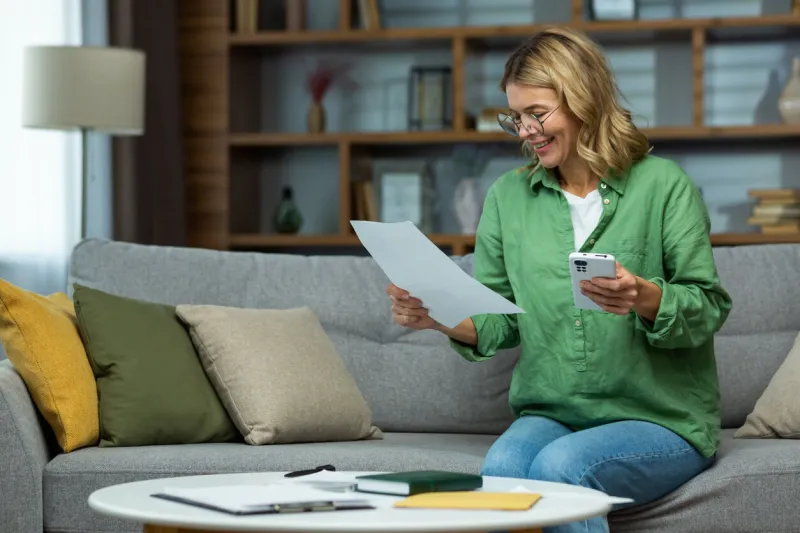 an elderly smilling woman sits at home on the sofa, holds the phone in her hands, works with documents, financial accounts, rent, loans, agreements