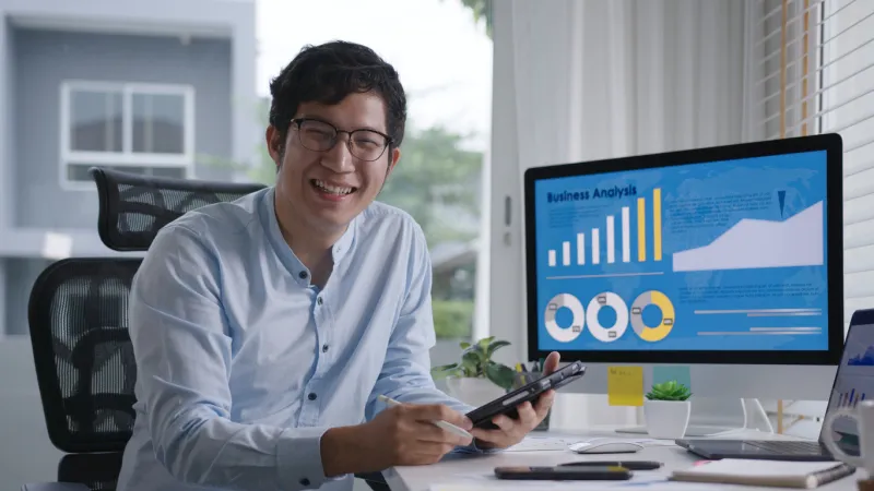 portrait head shot of young attractive asian man sitting smiling work multiple screen computer and smart tablet on table desk at home in concept freelance data analyst, data scientist for business