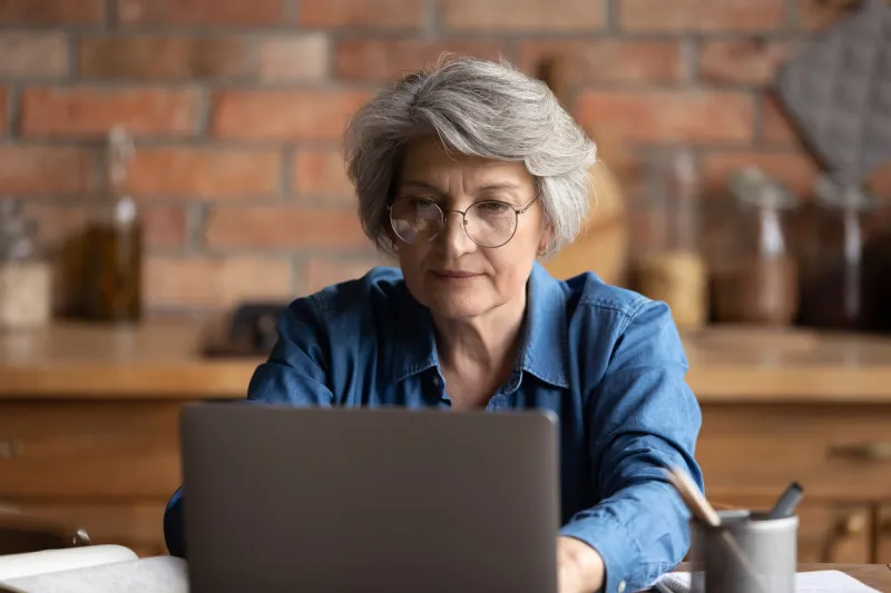 serious focused middle-aged woman in glasses working on laptop seated at table in kitchen, lead correspondence to client remotely, gain new knowledge use on-line resource, study on retirement concept