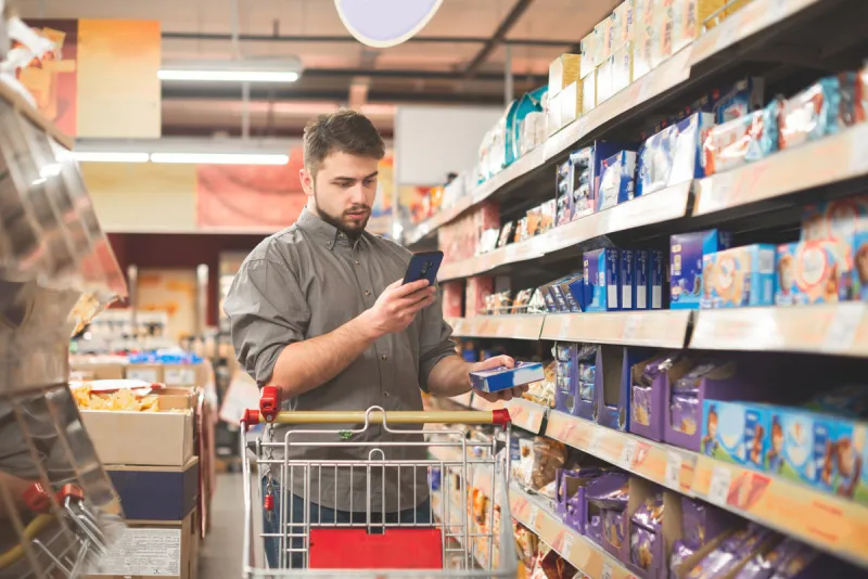 portrait of a man standing with a cart in the department of sweets in a supermarket, using a smartphone buyer with a cookie in his hands stands at the shelves and uses the internet on a smartphone
