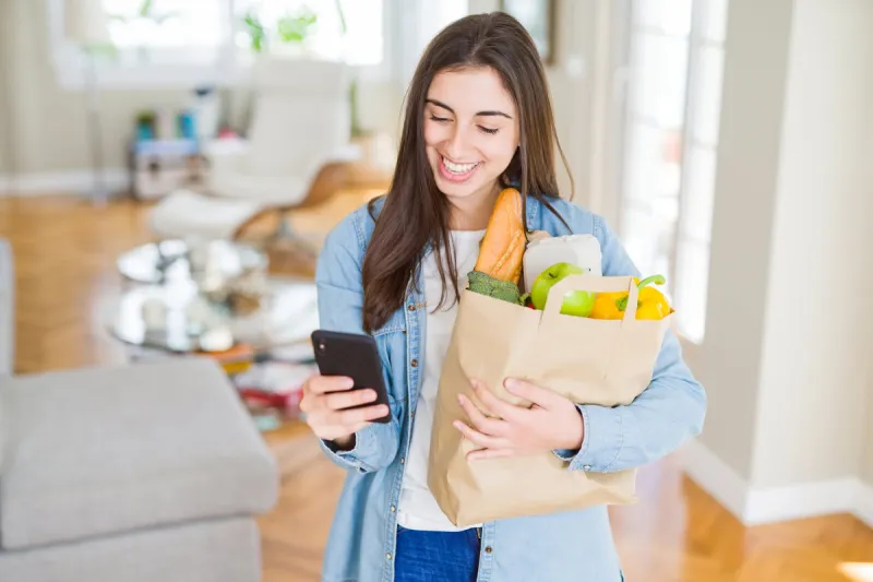 young woman holding a paper bag full of fresh groceries and using smartphone app for supermarket delivery
