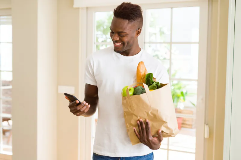 african man holding a paper bag full of groceries and using smarpthone buying online using app smiling