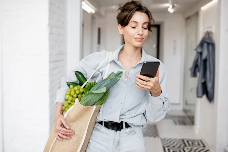 portrait of a young and cheerful woman standing with mobile phone and shopping bag full of fresh food at home concept of buying products online