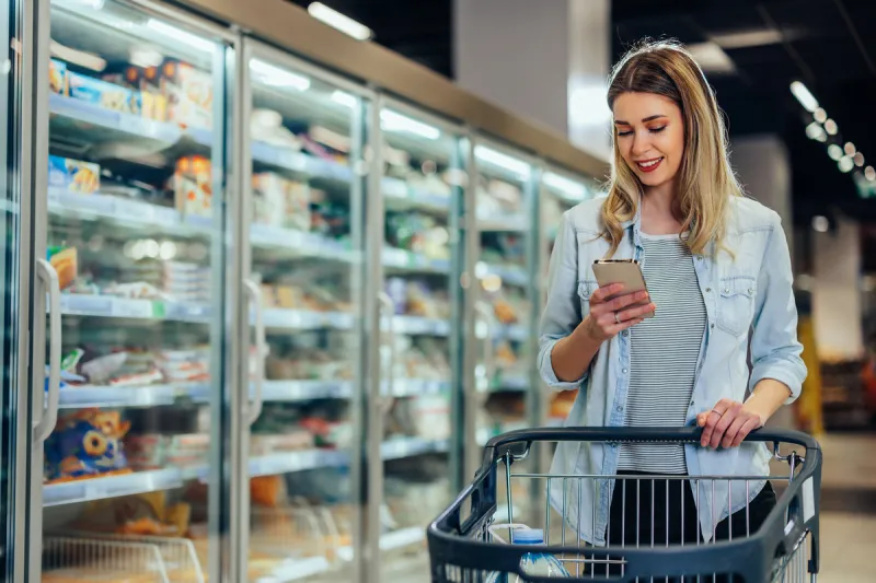 adult woman in grocery store pushing shopping trolley and using phone, reading shopping lists in app format