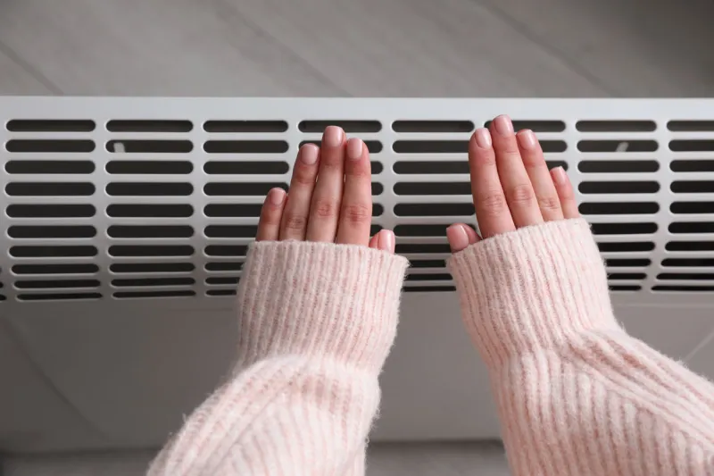 woman warming hands near electric heater at home, top view