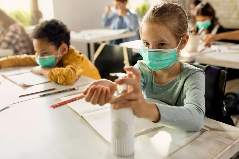 elementary student wearing protective face mask and disinfecting her hands in the classroom