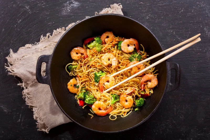 stir fried noodles with shrimps and vegetables in a wok on dark table, top view